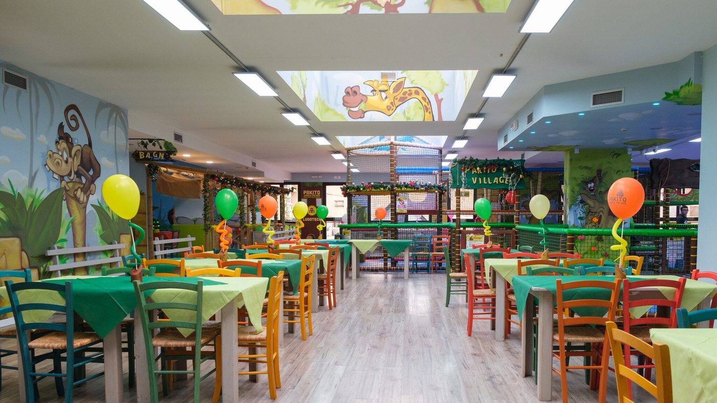 Sala interna del Pakito Jungle Palace Castelvetrano: ambiente jungle con murale giraffa e scimmia al soffitto, tavoli colorati verdi, insegna Pakito Village, rete di arrampicata sullo sfondo